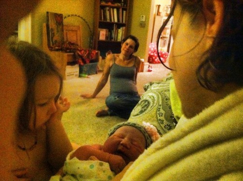 Melanie Moore, background, looks on at a new mother, baby, and family after a birth.