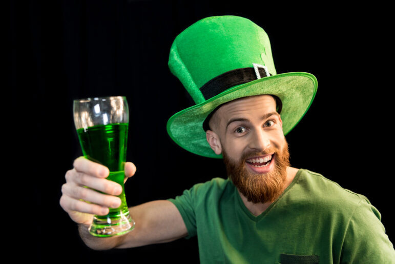 A man wearing a grene shirt and green hat and holding a glass of green beer, celebrating St.Patrick's day. 